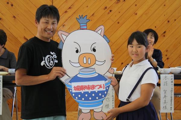 城東味まつりと書かれキャラクターが載ったカードを持つ小学生と男性の写真